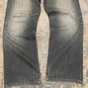 Pode incluir: Cal&ccedil;as de ganga cinza desbotadas com um design cl&aacute;ssico de cinco bolsos. As cal&ccedil;as de ganga t&ecirc;m um corte de perna reto e mostram sinais de desgaste, com &aacute;reas mais claras nas coxas e joelhos. O tecido tem uma apar&ecirc;ncia ligeiramente desgastada.