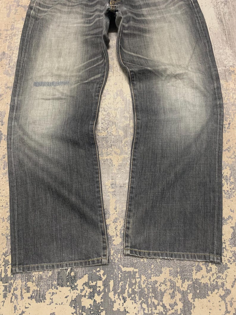 Pode incluir: Cal&ccedil;as de ganga cinza desbotadas com um design cl&aacute;ssico de cinco bolsos. As cal&ccedil;as de ganga t&ecirc;m um corte de perna reto e mostram sinais de desgaste, com &aacute;reas mais claras nas coxas e joelhos. O tecido tem uma apar&ecirc;ncia ligeiramente desgastada.