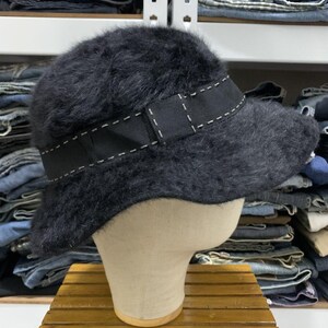 May include: A black, fuzzy bucket hat with a black ribbon band featuring white stitching. The hat is displayed on a mannequin head, showcasing its shape and texture. The hat is a stylish accessory.