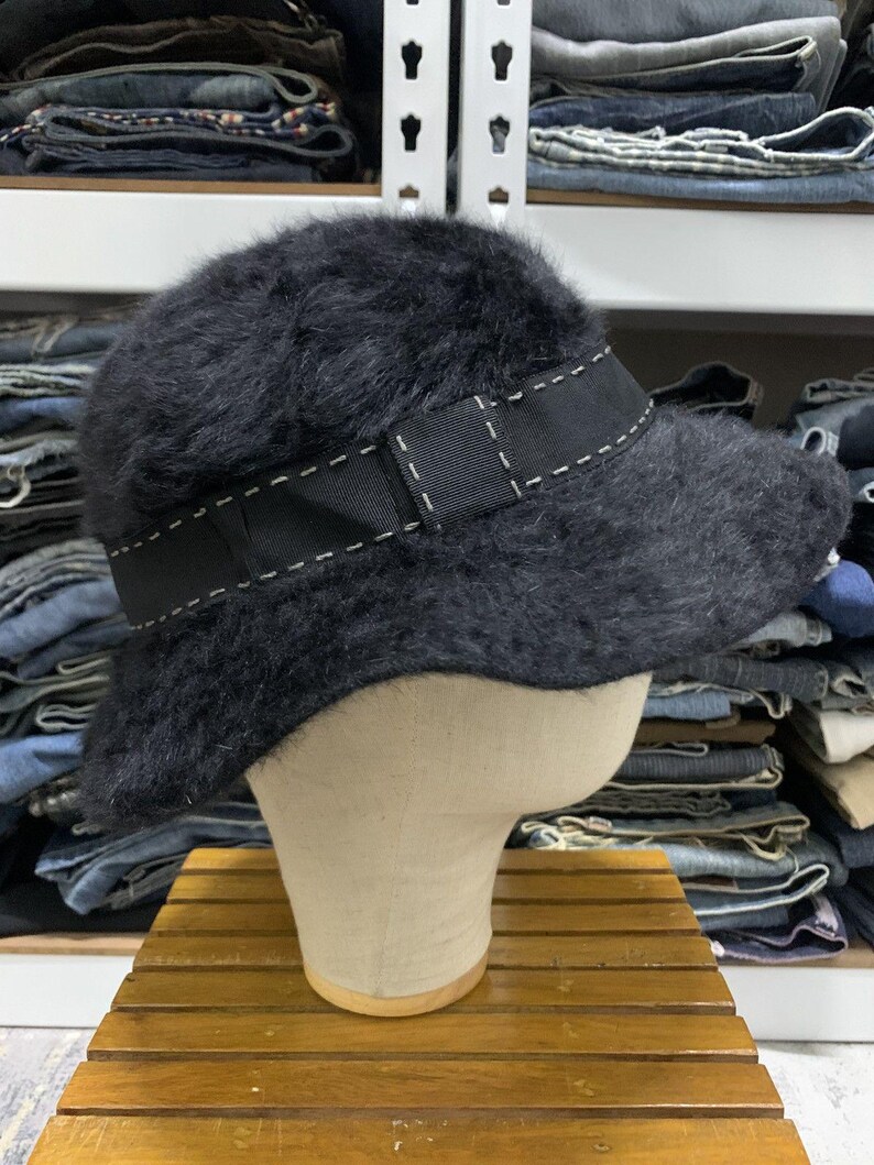 May include: A black, fuzzy bucket hat with a black ribbon band featuring white stitching. The hat is displayed on a mannequin head, showcasing its shape and texture. The hat is a stylish accessory.