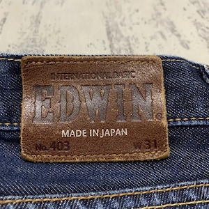 May include: Close-up of a brown leather patch on blue denim jeans. The patch reads "INTERNATIONAL BASIC EDWIN MADE IN JAPAN" with "No. 403" and "W 31" printed on it. The jeans have yellow stitching.
