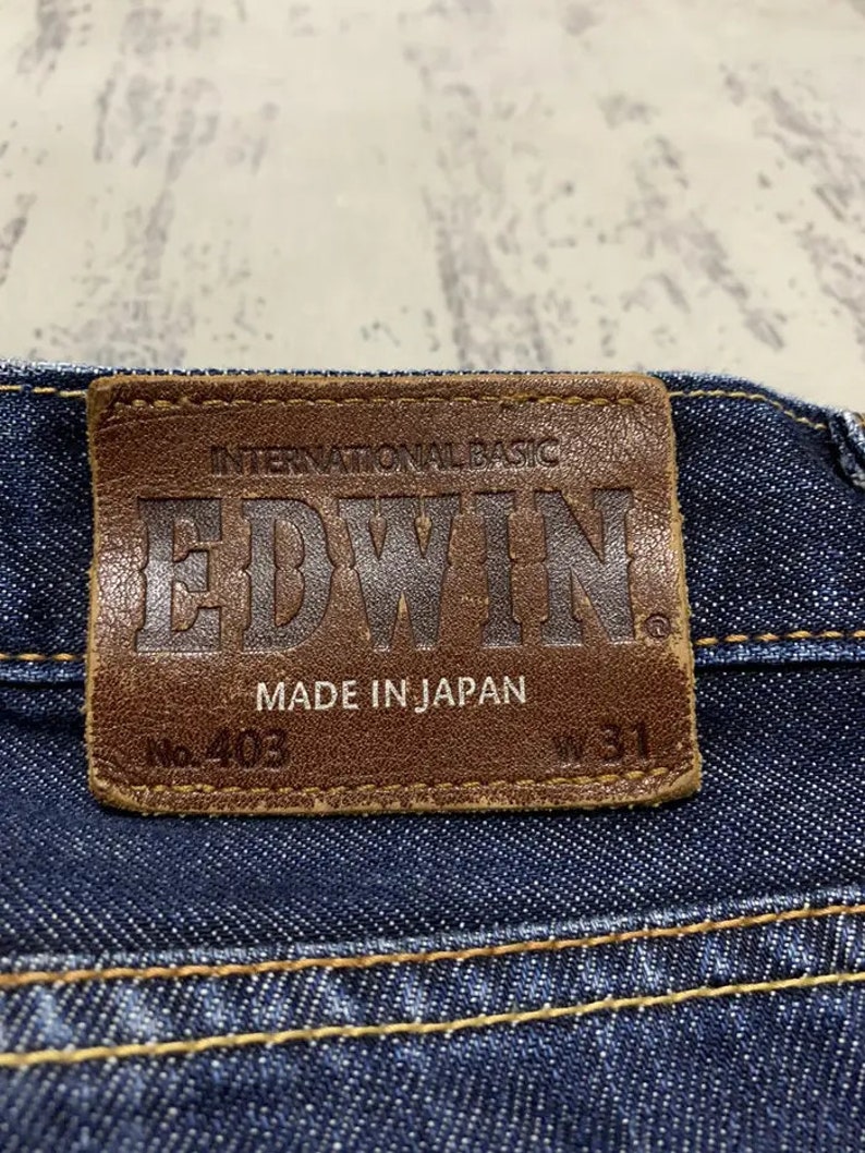 May include: Close-up of a brown leather patch on blue denim jeans. The patch reads "INTERNATIONAL BASIC EDWIN MADE IN JAPAN" with "No. 403" and "W 31" printed on it. The jeans have yellow stitching.