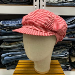 May include: A newsboy cap featuring a vibrant red and white vertical pinstripe pattern. The hat has a rounded, multi-paneled crown and a stiff, short brim extending from the front, displayed on a neutral-colored mannequin head form.