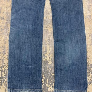 May include: A pair of blue denim jeans. The jeans have a classic five-pocket design, a straight leg, and a medium wash. The fabric shows subtle fading and wear, suggesting a comfortable fit. The jeans are laid flat on a textured surface.