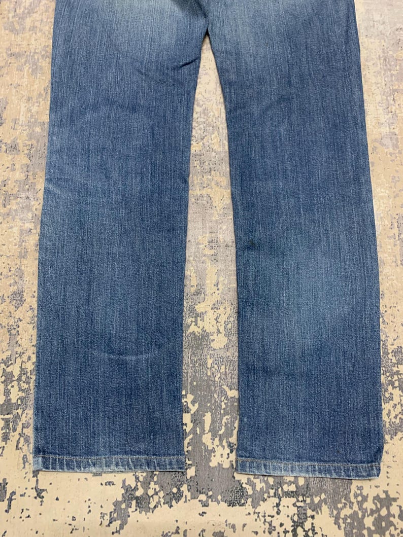 May include: A pair of blue denim jeans. The jeans have a classic five-pocket design, a straight leg, and a medium wash. The fabric shows subtle fading and wear, suggesting a comfortable fit. The jeans are laid flat on a textured surface.