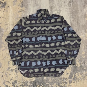 Può includere: Una camicia a maniche lunghe con un motivo unico. La camicia presenta un colore di base blu scuro con forme ondulate e irregolari in tonalità di grigio e azzurro. Ha un colletto classico e due tasche sul petto.
