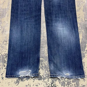 May include: Close-up of a pair of blue denim jeans. The jeans have a faded wash and distressed details at the hem. The fabric shows wear and tear, with visible fraying and small holes near the bottom.