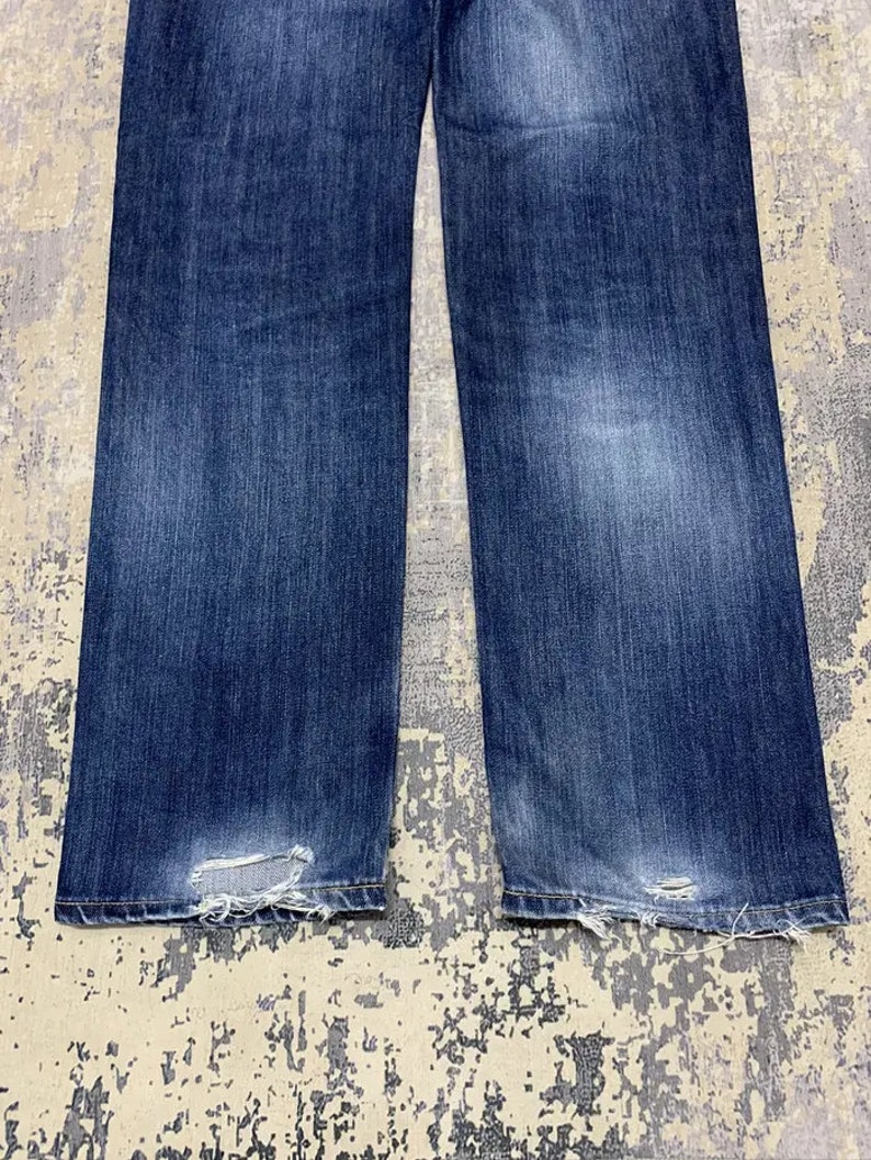 May include: Close-up of a pair of blue denim jeans. The jeans have a faded wash and distressed details at the hem. The fabric shows wear and tear, with visible fraying and small holes near the bottom.
