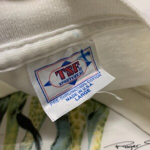 Puede incluir: Camiseta blanca con estampado de jirafas y etiqueta que dice "TSF Sportswear" y "Made in U.S.A.". La camiseta es pre-encogida, 100% algod&oacute;n y talla Large. El estampado de jirafas es visible.
