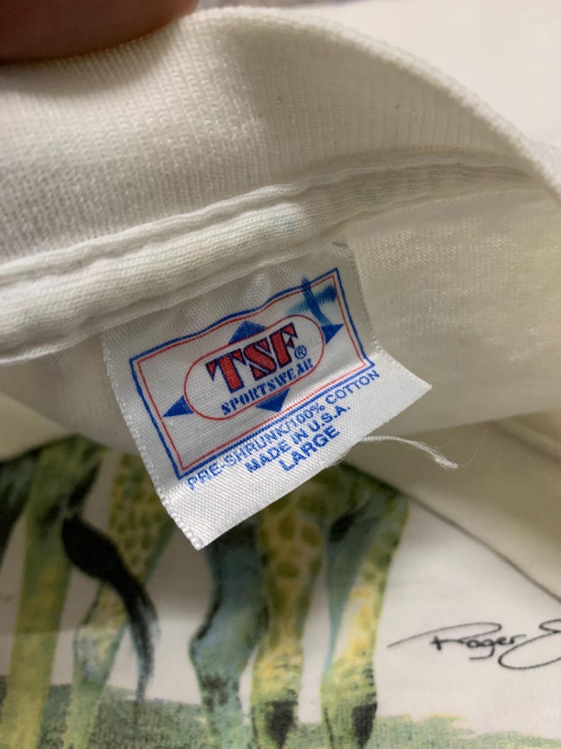 Puede incluir: Camiseta blanca con estampado de jirafas y etiqueta que dice "TSF Sportswear" y "Made in U.S.A.". La camiseta es pre-encogida, 100% algod&oacute;n y talla Large. El estampado de jirafas es visible.