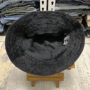 May include: A black, fuzzy bucket hat. The hat is made of a soft, textured material and has a dark interior. The hat is sitting on a small wooden stand. The hat's brim is slightly curved.