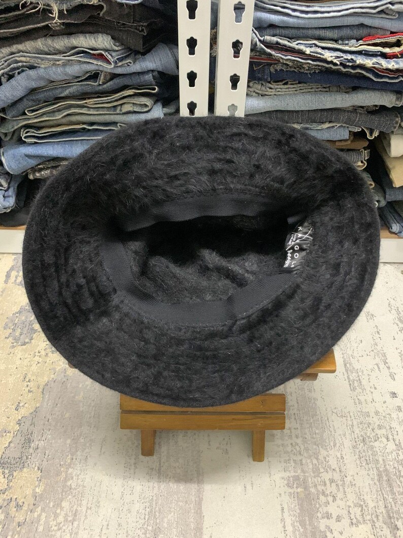 May include: A black, fuzzy bucket hat. The hat is made of a soft, textured material and has a dark interior. The hat is sitting on a small wooden stand. The hat's brim is slightly curved.