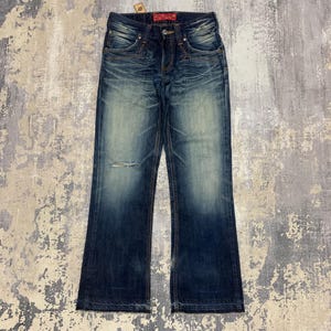 May include: Dark wash denim jeans with a distressed look. The jeans have a faded appearance, a small rip on the left knee, and a red tag on the back. The jeans have a classic five-pocket design and a button and zipper closure.