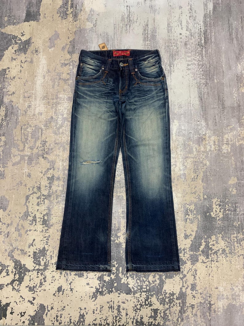 May include: Dark wash denim jeans with a distressed look. The jeans have a faded appearance, a small rip on the left knee, and a red tag on the back. The jeans have a classic five-pocket design and a button and zipper closure.