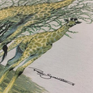 Puede incluir: Camiseta blanca con una ilustraci&oacute;n detallada de dos jirafas con un patr&oacute;n de manchas verdes y amarillas, sobre un fondo de follaje verde. La firma del artista, "Roger Swainston", es visible en escritura negra.