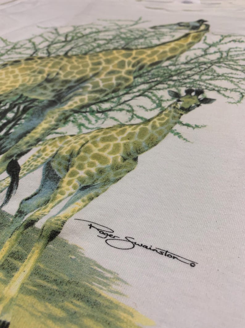 Puede incluir: Camiseta blanca con una ilustraci&oacute;n detallada de dos jirafas con un patr&oacute;n de manchas verdes y amarillas, sobre un fondo de follaje verde. La firma del artista, "Roger Swainston", es visible en escritura negra.