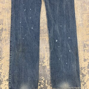 May include: Faded blue denim jeans with a straight leg cut. The jeans have white paint splatters throughout the front. The denim shows signs of wear and fading, particularly around the knees and hem.