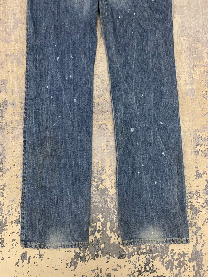 May include: Faded blue denim jeans with a straight leg cut. The jeans have white paint splatters throughout the front. The denim shows signs of wear and fading, particularly around the knees and hem.