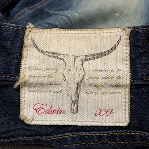 May include: Close-up of a denim jeans label. The label is rectangular with a skull graphic and text that reads "Edwin". The label is stitched onto the dark blue denim fabric. The text on the label is in red and black.