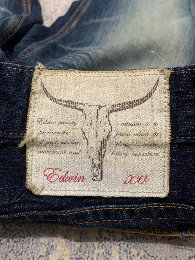 May include: Close-up of a denim jeans label. The label is rectangular with a skull graphic and text that reads "Edwin". The label is stitched onto the dark blue denim fabric. The text on the label is in red and black.