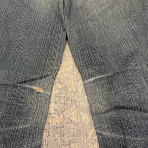 Op de afbeelding: Verbleekte blauwe denim jeans met zichtbare slijtage. De jeans hebben een klassiek ontwerp met vijf zakken, een knoop- en ritssluiting en een versleten plek op het linkerbeen. De achterzakken hebben decoratieve stiksels.