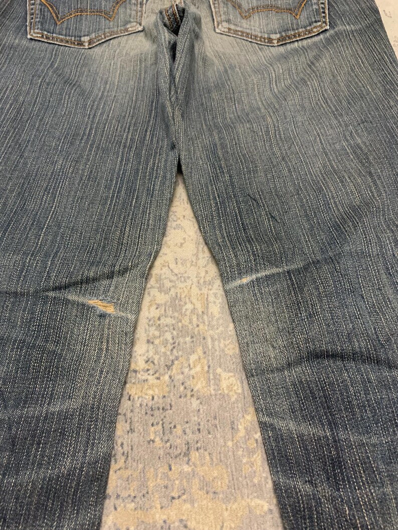 Op de afbeelding: Verbleekte blauwe denim jeans met zichtbare slijtage. De jeans hebben een klassiek ontwerp met vijf zakken, een knoop- en ritssluiting en een versleten plek op het linkerbeen. De achterzakken hebben decoratieve stiksels.
