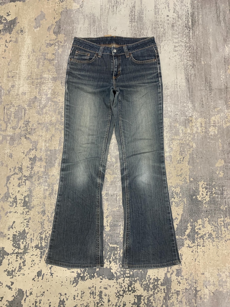 Op de afbeelding: Blauwe spijkerbroek met een klassiek ontwerp met vijf zakken en een lichte flare aan de zoom. De jeans heeft een vervaagde wassing en een knoop- en ritssluiting. De stof lijkt versleten, wat wijst op een vintage of tweedehands item.