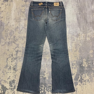 Op de afbeelding: Blauwe jeans van denim met een uitlopende pijp. De jeans hebben twee achterzakken, een klein muntzakje en een leren label met de merknaam "EDWIN". De jeans liggen plat op een getextureerd oppervlak.