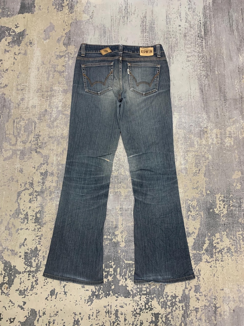 Op de afbeelding: Blauwe jeans van denim met een uitlopende pijp. De jeans hebben twee achterzakken, een klein muntzakje en een leren label met de merknaam "EDWIN". De jeans liggen plat op een getextureerd oppervlak.