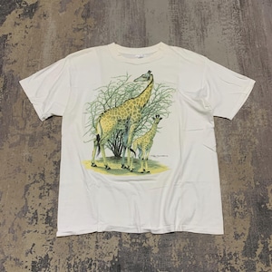Puede incluir: Camiseta blanca con un gr&aacute;fico de dos jirafas, una adulta y una cr&iacute;a, frente a un arbusto verde. Las jirafas tienen un patr&oacute;n marr&oacute;n y amarillo. La camiseta est&aacute; hecha de una tela blanca suave.