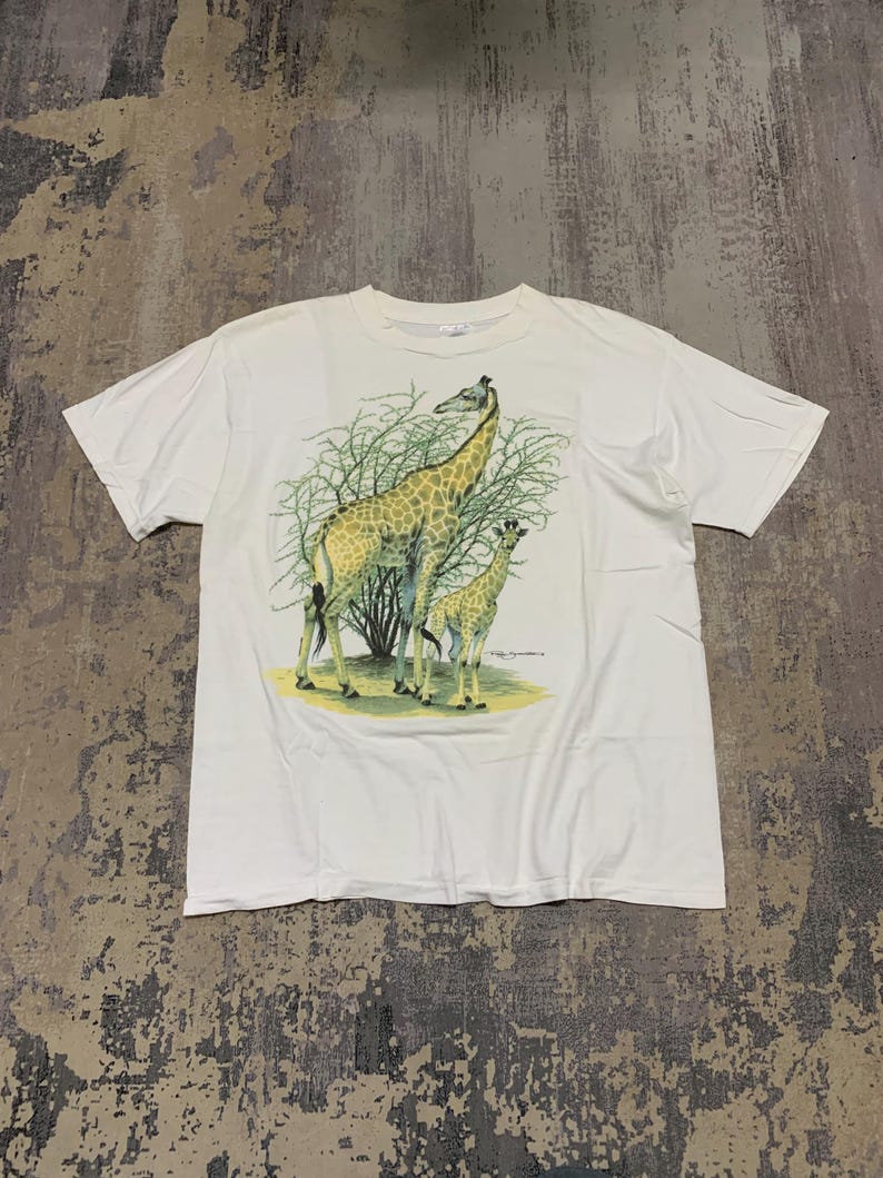 Puede incluir: Camiseta blanca con un gr&aacute;fico de dos jirafas, una adulta y una cr&iacute;a, frente a un arbusto verde. Las jirafas tienen un patr&oacute;n marr&oacute;n y amarillo. La camiseta est&aacute; hecha de una tela blanca suave.
