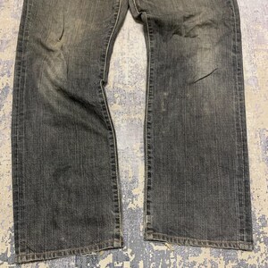 Pu&ograve; includere: Jeans di denim neri sbiaditi con taglio dritto. I jeans mostrano segni di usura con macchie pi&ugrave; chiare e sottili sbiaditure. Il tessuto ha un aspetto leggermente usurato, con linee verticali visibili. I jeans sono stesi piatti, mostrando la parte anteriore del capo.