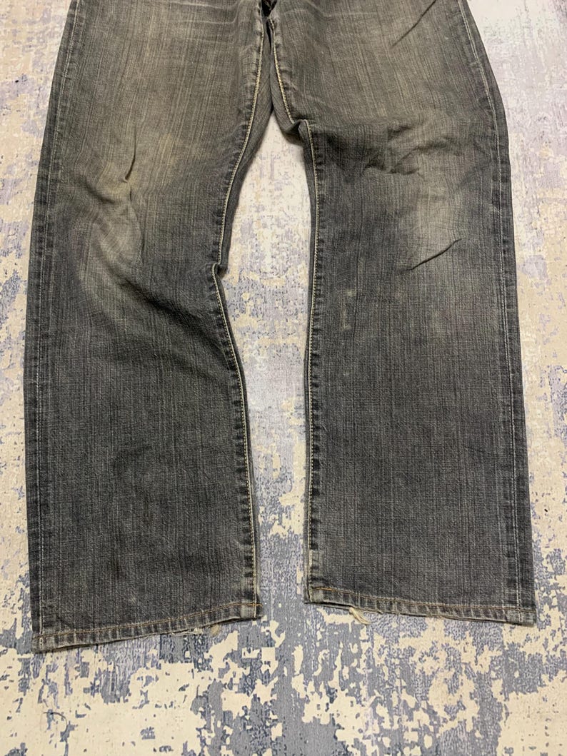 Pu&ograve; includere: Jeans di denim neri sbiaditi con taglio dritto. I jeans mostrano segni di usura con macchie pi&ugrave; chiare e sottili sbiaditure. Il tessuto ha un aspetto leggermente usurato, con linee verticali visibili. I jeans sono stesi piatti, mostrando la parte anteriore del capo.
