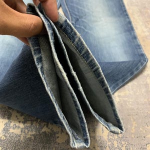 May include: Close-up of folded blue denim jeans. The jeans have a classic five-pocket design and a faded wash. The denim is a medium weight, and the seams are neatly stitched. The jeans are folded to show the inside of the hem.