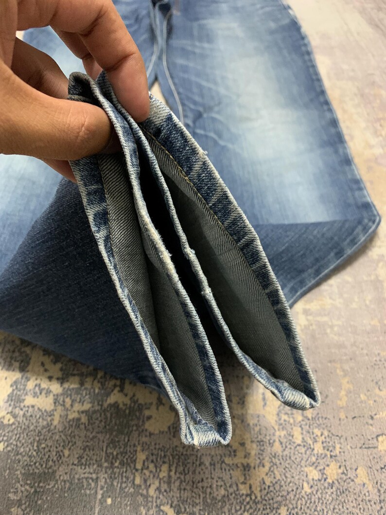 May include: Close-up of folded blue denim jeans. The jeans have a classic five-pocket design and a faded wash. The denim is a medium weight, and the seams are neatly stitched. The jeans are folded to show the inside of the hem.