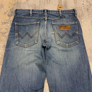 May include: Light blue denim jeans with classic five-pocket design. Features a brown leather patch with the "Wrangler" logo on the back pocket. The jeans have a faded wash and a straight leg cut.