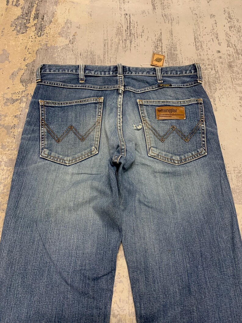 May include: Light blue denim jeans with classic five-pocket design. Features a brown leather patch with the "Wrangler" logo on the back pocket. The jeans have a faded wash and a straight leg cut.