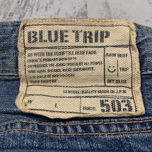May include: Close-up of a denim label with the text "BLUE TRIP" in black. The label is rectangular with a beige background and black text. Additional text includes "RAW BLUE", "DRY BLUE", and "503".