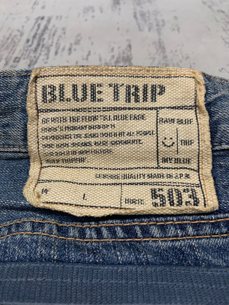 May include: Close-up of a denim label with the text "BLUE TRIP" in black. The label is rectangular with a beige background and black text. Additional text includes "RAW BLUE", "DRY BLUE", and "503".