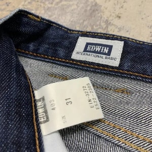 May include: Close-up of dark blue denim jeans with the brand label "EDWIN INTERNATIONAL BASIC" visible. A white tag displays "SIZE 31" and other product information. The jeans have visible stitching.