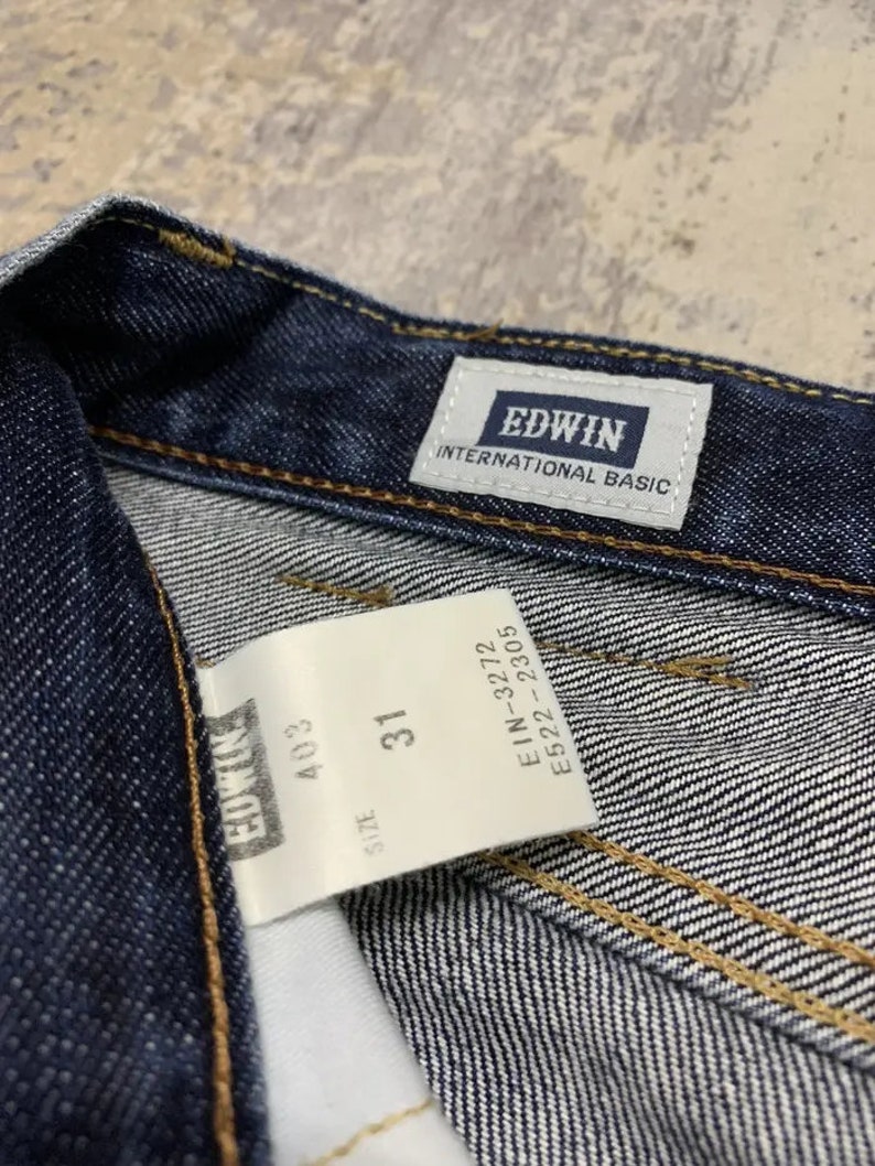 May include: Close-up of dark blue denim jeans with the brand label "EDWIN INTERNATIONAL BASIC" visible. A white tag displays "SIZE 31" and other product information. The jeans have visible stitching.