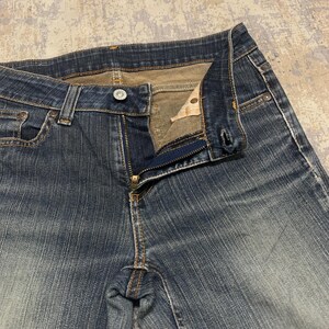 Op de afbeelding: Close-up van een blauwe denim jeans met een gedeeltelijk open rits. De jeans heeft een knoopsluiting en een klassiek vijf-zakken ontwerp. De denim toont vervaging en slijtage, met zichtbare stikseldetails.