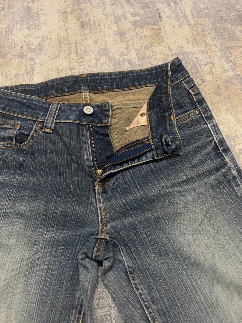 Op de afbeelding: Close-up van een blauwe denim jeans met een gedeeltelijk open rits. De jeans heeft een knoopsluiting en een klassiek vijf-zakken ontwerp. De denim toont vervaging en slijtage, met zichtbare stikseldetails.