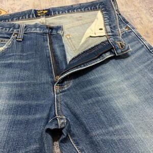 May include: Close-up of blue denim jeans with a partially unzipped fly. The jeans have a classic five-pocket design, with visible stitching and a Wrangler label at the waistband. The denim has a faded, worn appearance.