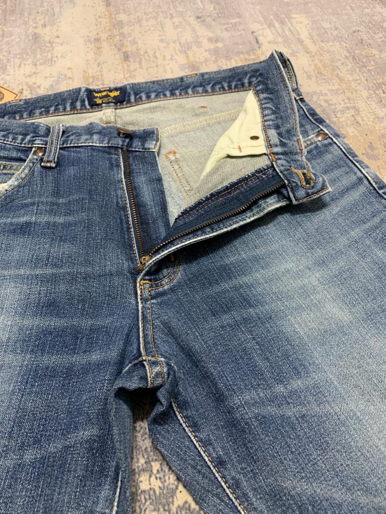 May include: Close-up of blue denim jeans with a partially unzipped fly. The jeans have a classic five-pocket design, with visible stitching and a Wrangler label at the waistband. The denim has a faded, worn appearance.