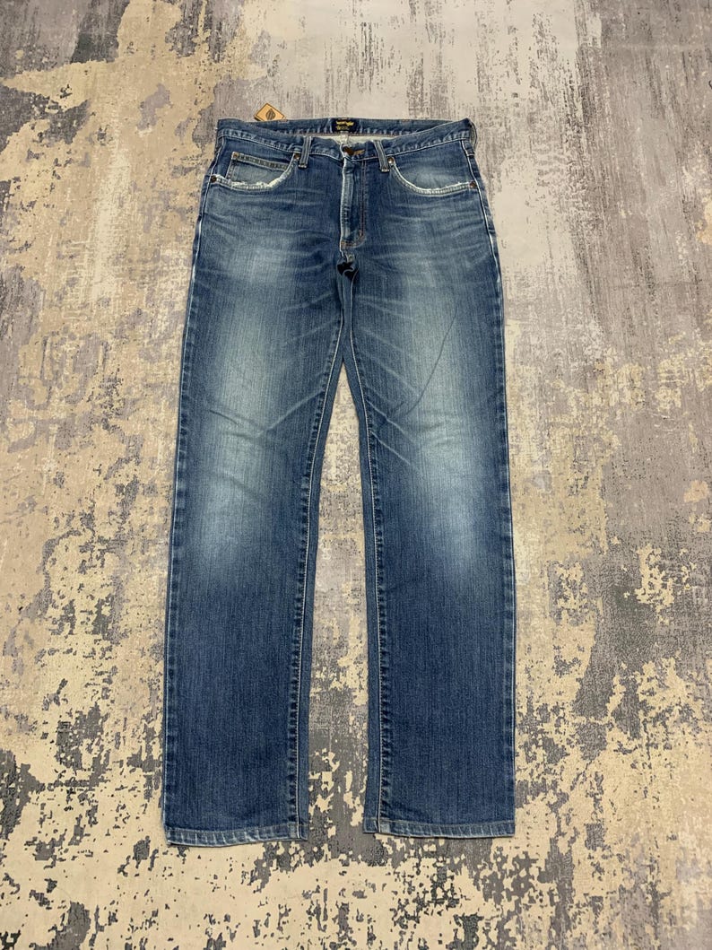May include: Blue denim jeans with a faded wash and a classic five-pocket design. The jeans have a straight leg cut and a button and zipper closure. The Wrangler brand name is visible on the back pocket.