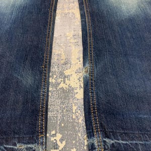May include: Close-up of blue denim jeans with distressed details. The jeans have visible wear and tear, with frayed edges and a faded wash. The seams are highlighted with contrasting yellow stitching.