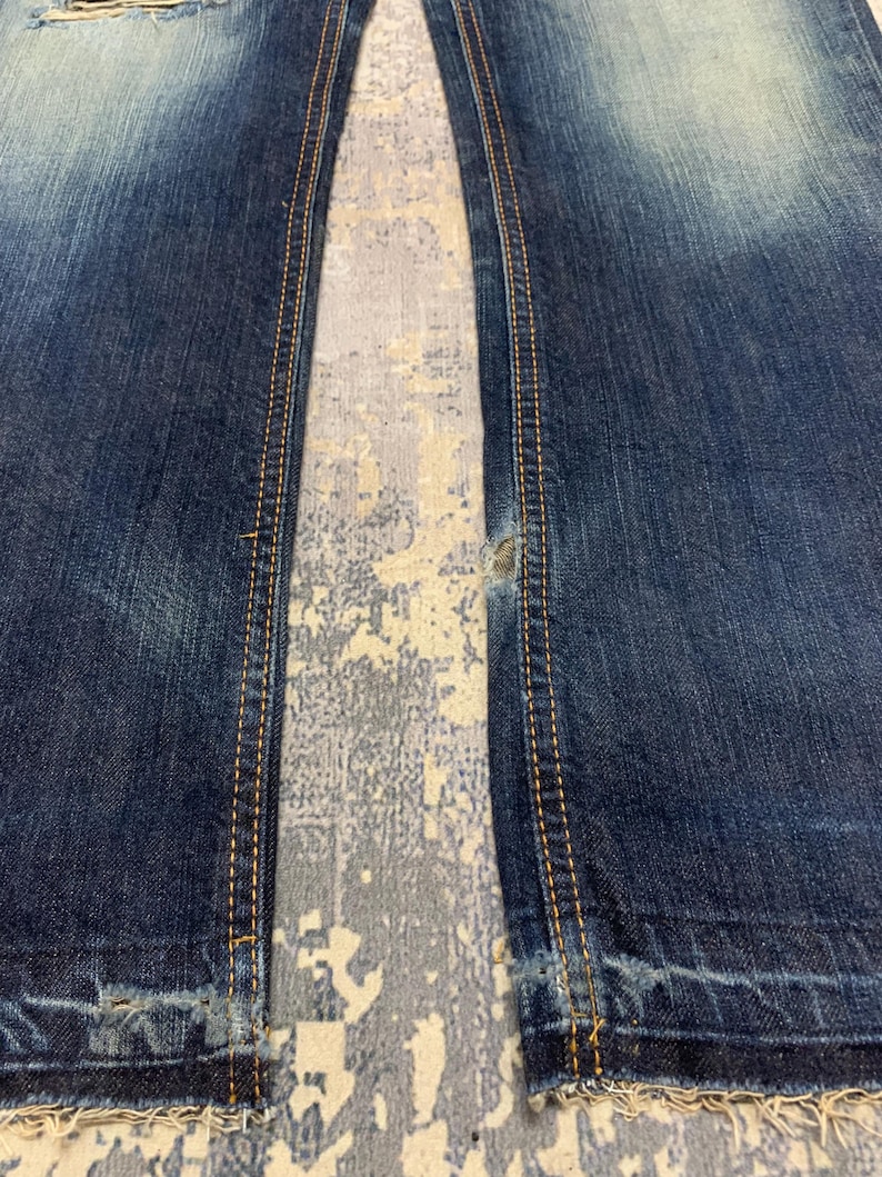 May include: Close-up of blue denim jeans with distressed details. The jeans have visible wear and tear, with frayed edges and a faded wash. The seams are highlighted with contrasting yellow stitching.