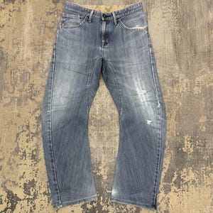 May include: Faded blue denim jeans with a classic five-pocket design. The jeans have a relaxed fit, with slight distressing on the front. The jeans have a button and zip closure. The jeans are laid flat on a textured surface.