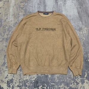 May include: A tan-colored sweatshirt with the text "U.P. Renoma UNIFORME PRESTIGE" in dark brown lettering. The sweatshirt has a crew neck and long sleeves. The collar and the inside of the neck are brown.